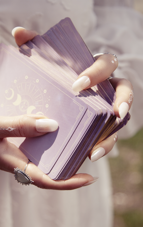 Zwei Hände mit hellen, länglichen Fingernägeln halten einen Stapel blasslila Karten mit sanften, hellen Symbolen darauf. Die Hände sind leicht verschwommen und der Hintergrund ist hell und unscharf.