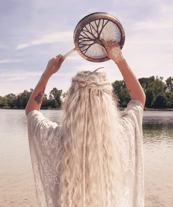 body-image-subpage Eine Frau mit langem blonden Haar steht am Ufer eines Sees und hält über ihrem Kopf eine Trommel.
