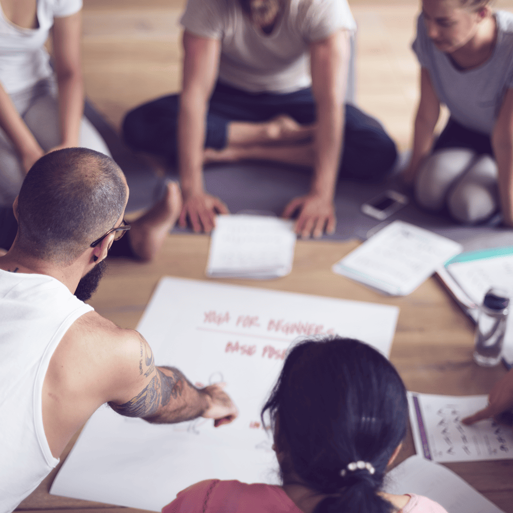 Eine Gruppe von Menschen sitzt auf dem Boden in einem Kreis. Vor ihnen liegen aufgeschlagene hefte und Notizen. Ein Mann links unten am Bildrand zeigt mit einer Hand auf ein großes Plakat, das in der Mitte des Sitzkreises liegt.